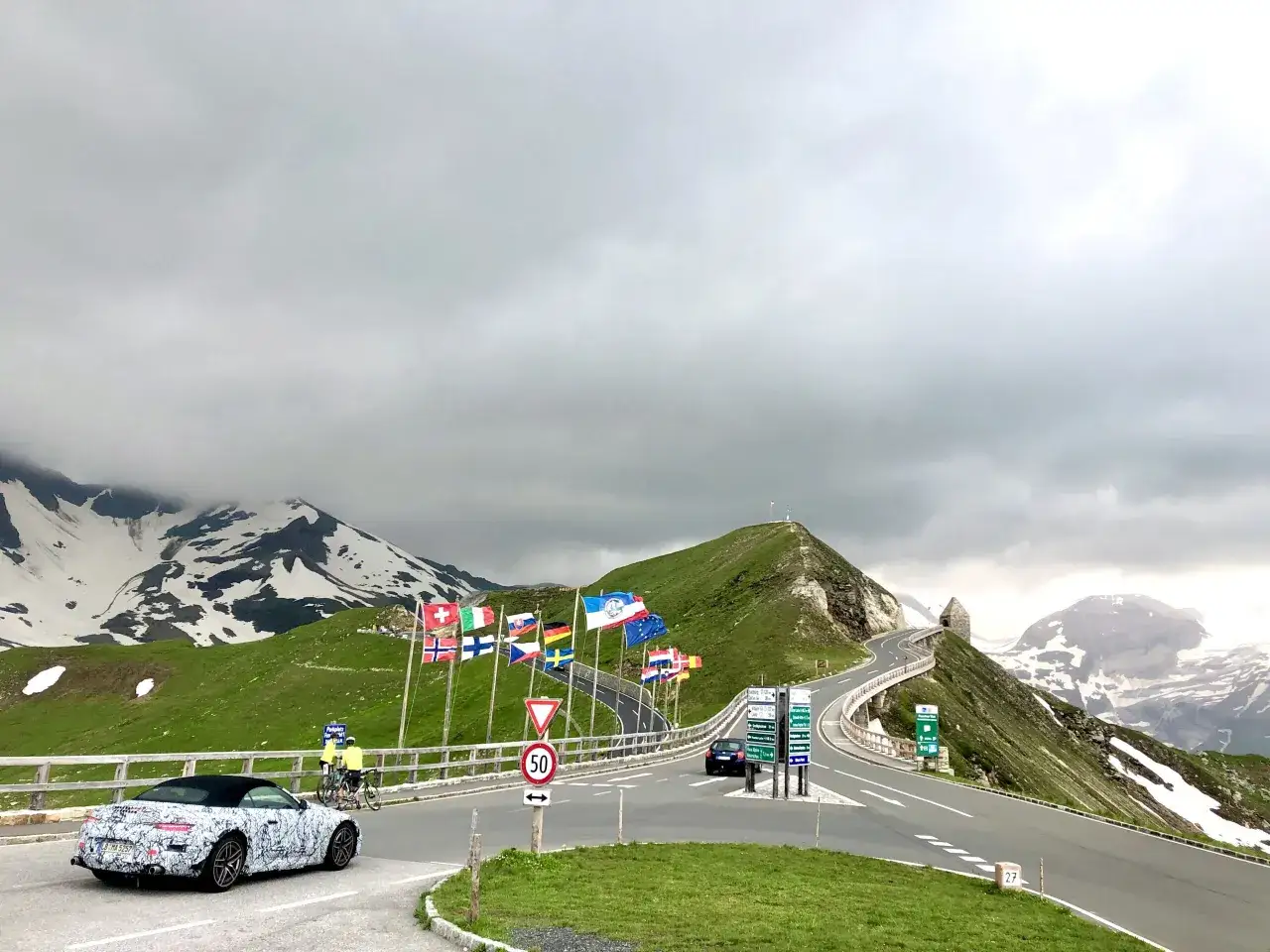 Wjazd na górską serpentynę, jedną z najpiękniejszych dróg w Europie, z widokiem na ośnieżone szczyty i flagi państw.
