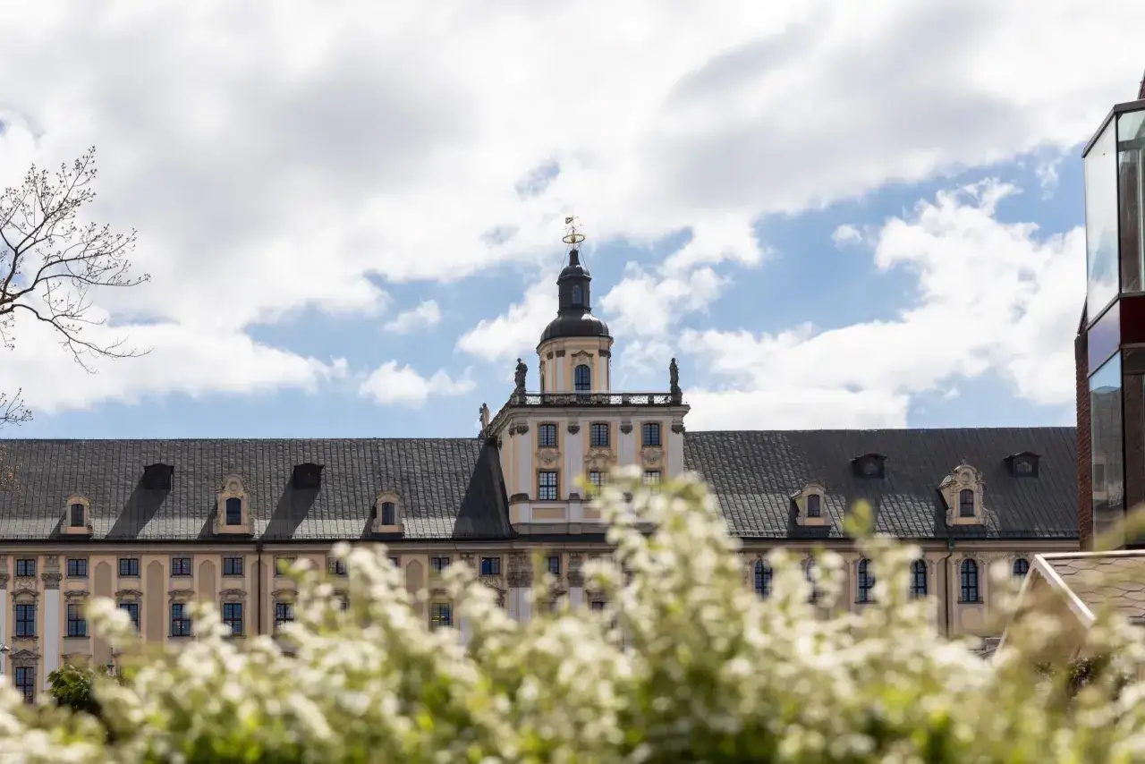 Gmach Uniwersytetu Wrocławskiego z wieżą, na tle błękitnego nieba z chmurami i kwitnącymi krzewami.