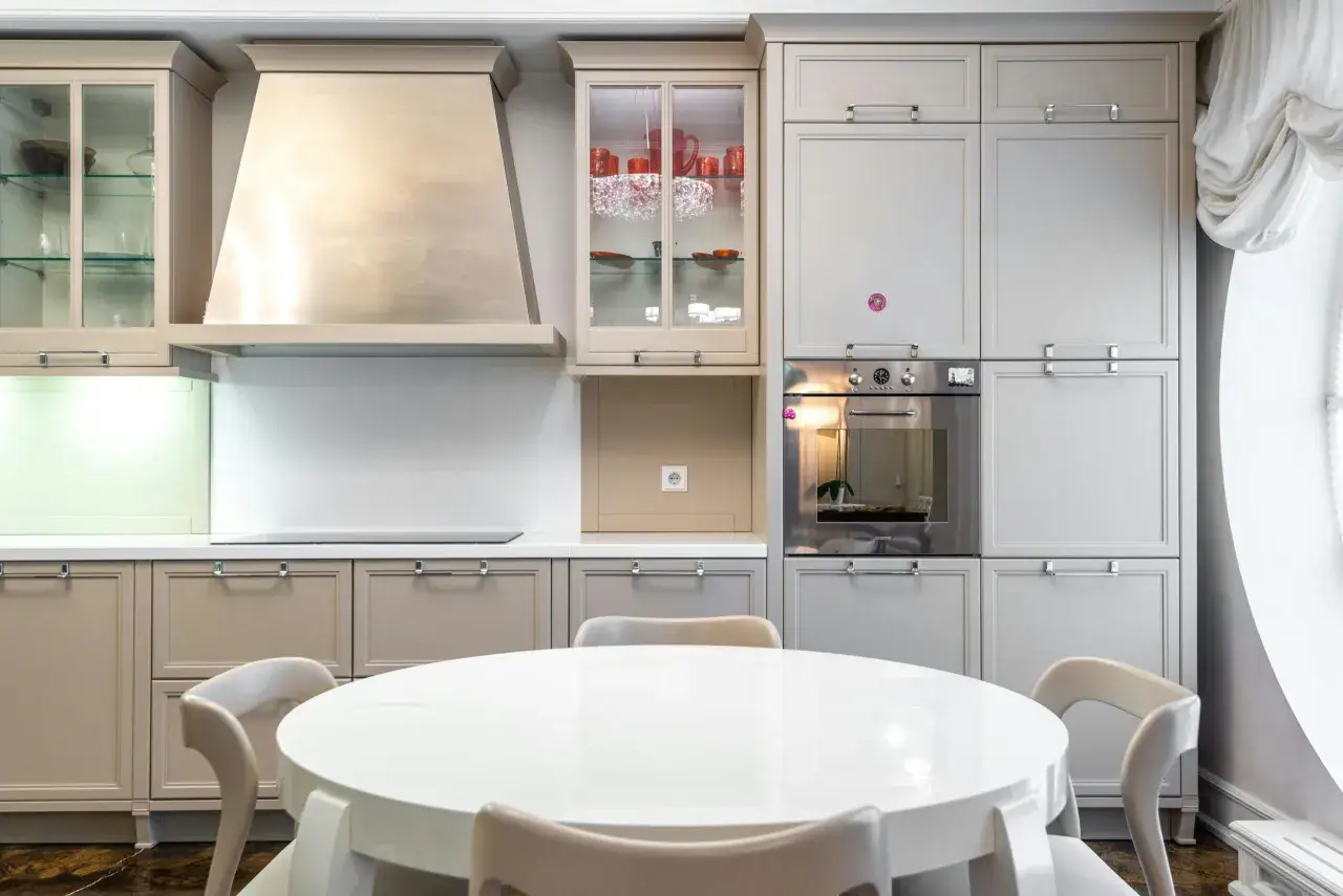 Cocina moderna con mesa redonda blanca y sillas. Gabinetes grises y beige, campana extractora de acero inoxidable. Ideal para ver catálogos de cocinas y precios.