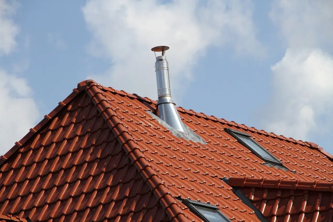 Metalowy komin na czerwonym, dachówkowym dachu pod błękitnym niebem z chmurami.