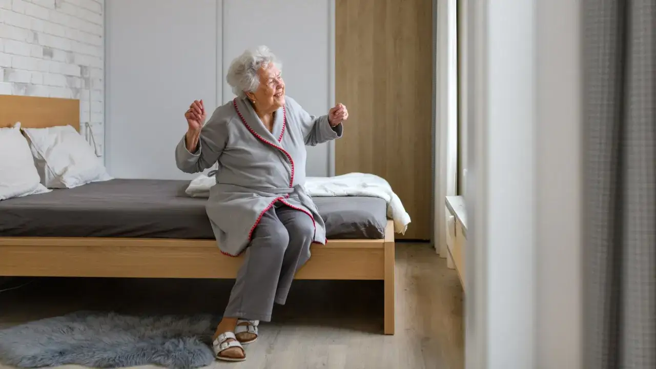 Uśmiechnięta starsza pani w szlafroku siedzi na łóżku, zastanawiając się, jaki wybrać pokój w sanatorium.