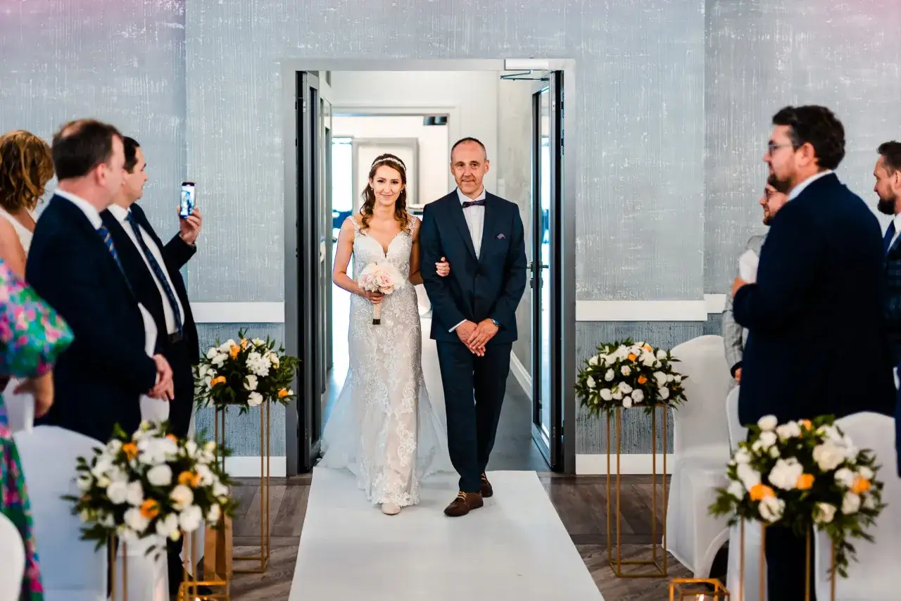 Panna młoda w sukni ślubnej idzie z ojcem do ołtarza, obok bukiety kwiatów.