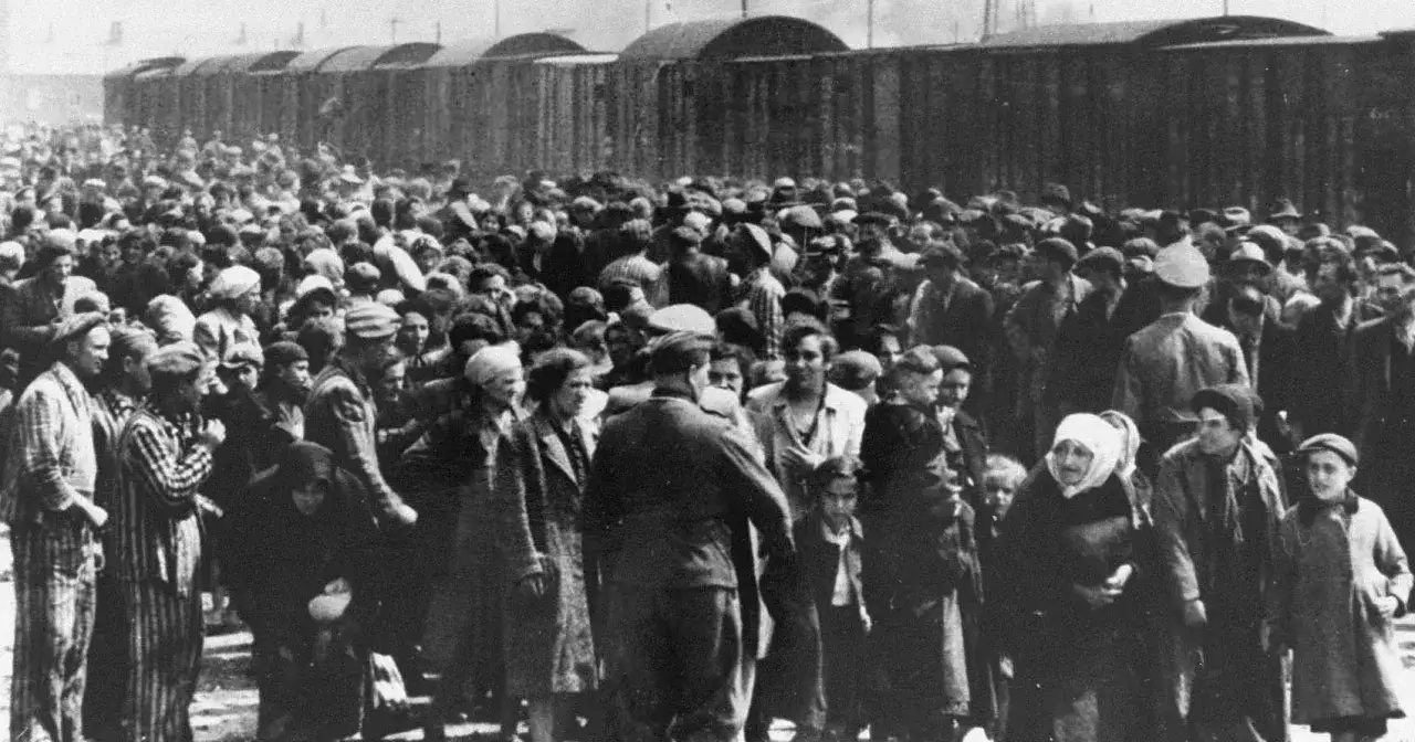Tłum ludzi, w tym dzieci, zgromadzony przy wagonach kolejowych. Każdy człowiek zlagrowany jako ofiara zbrodniczego systemu, pozbawiony godności i nadziei.
