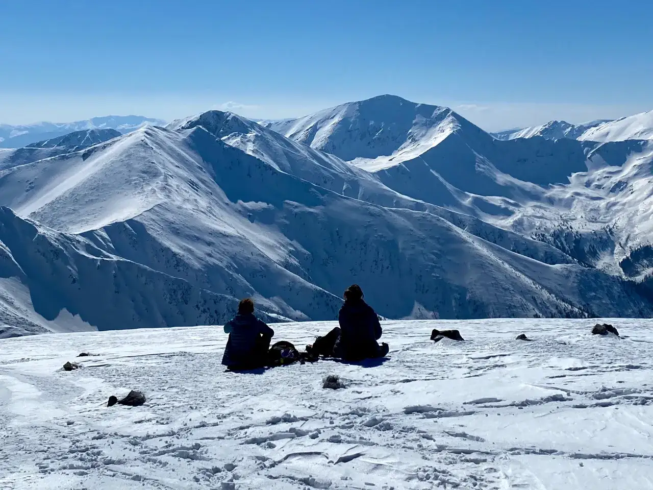 Dwie osoby odpoczywają na ośnieżonym szczycie, podziwiając majestatyczne Czerwone Wierchy. Czy szlak na Czerwone Wierchy jest trudny? Widok sugeruje, że warto podjąć wyzwanie.