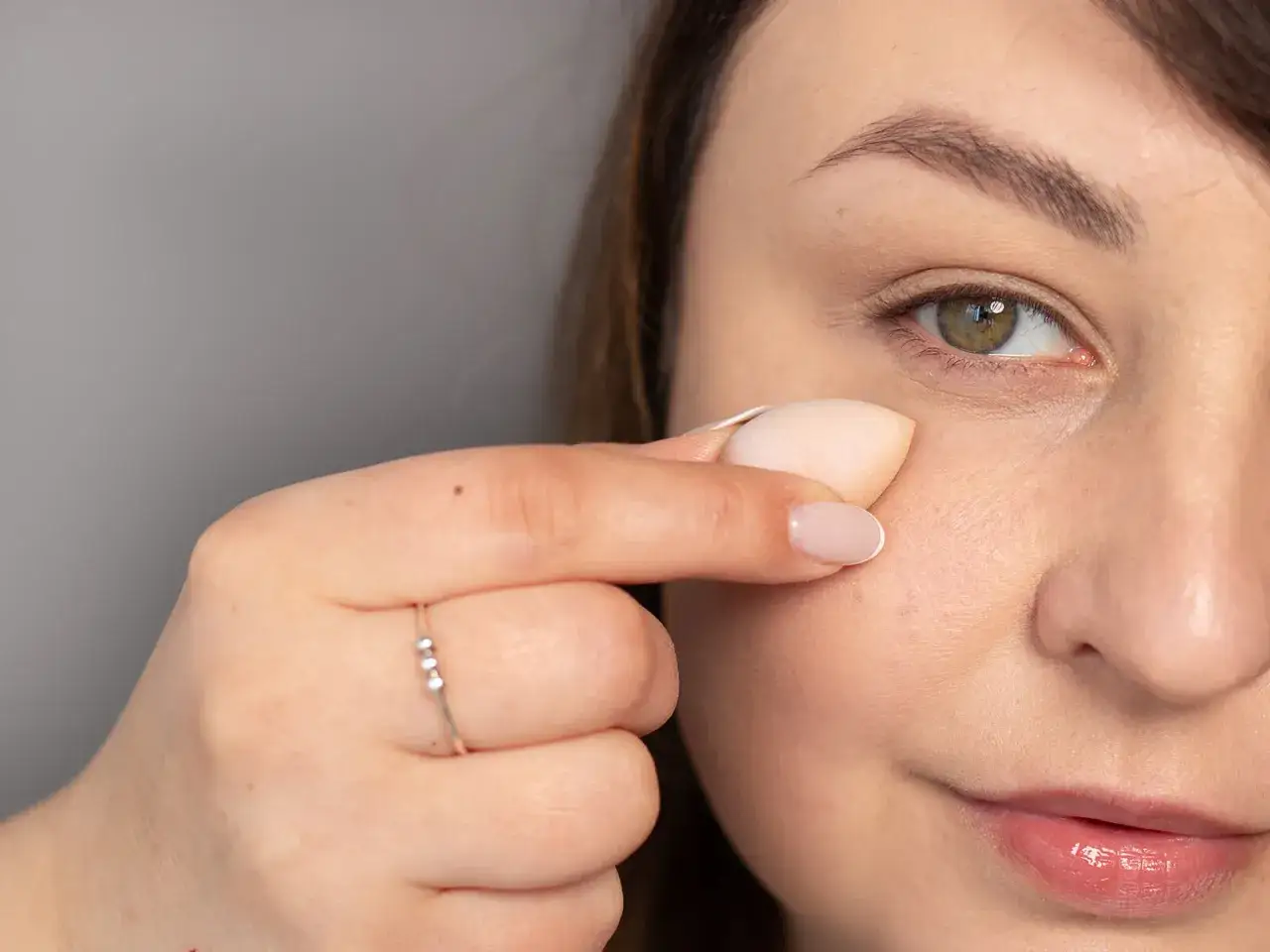 Dłoń z pierścionkiem aplikuje gąbeczką korektor pod oko.