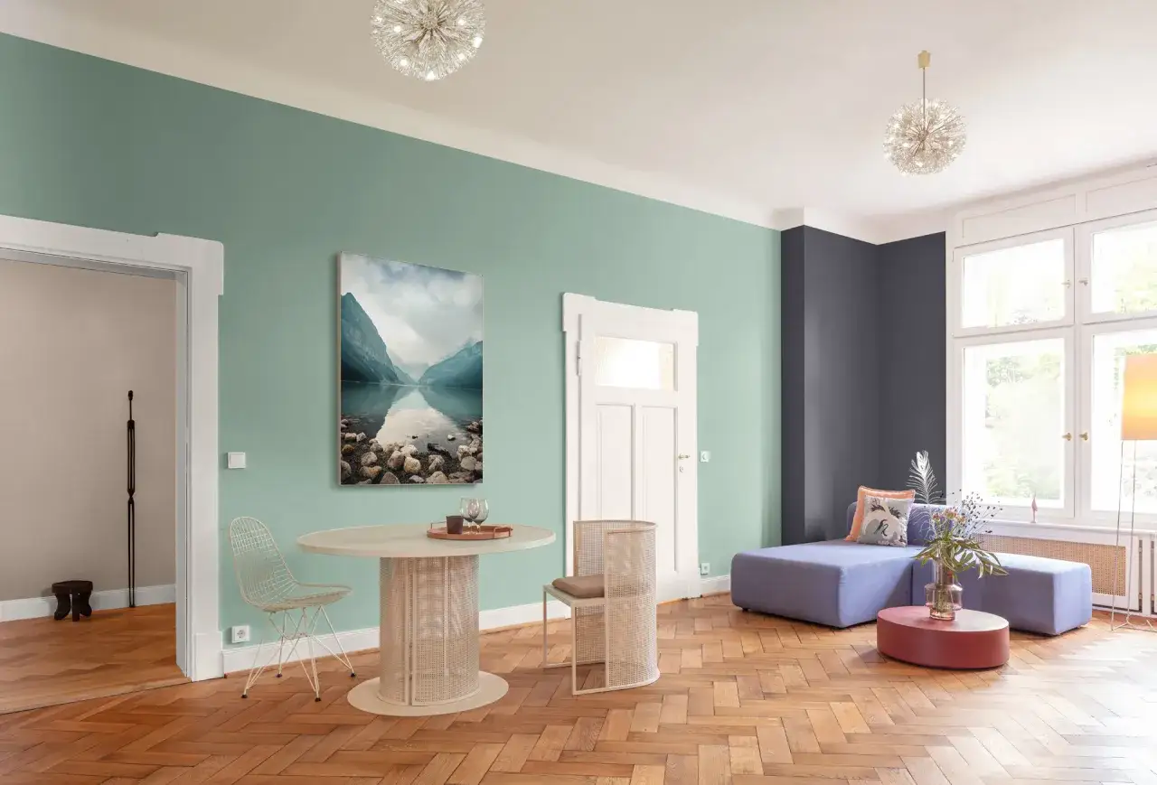 Nowoczesne wnętrze z miętową ścianą, obrazem górskiego jeziora, okrągłym stołem i fioletową kanapą.