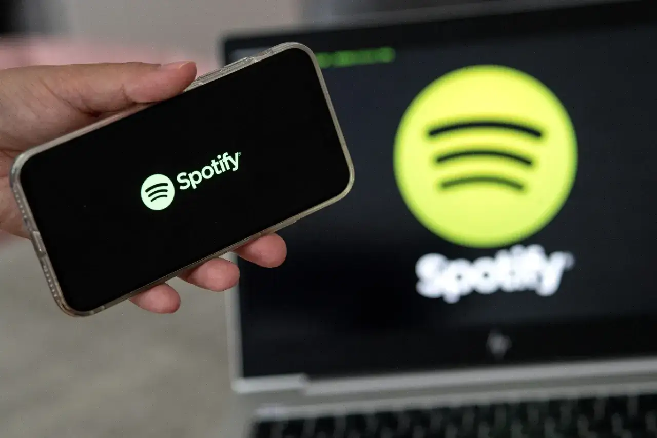 Dłoń trzyma smartfon z logo Spotify. W tle ekran laptopa z tym samym logo.