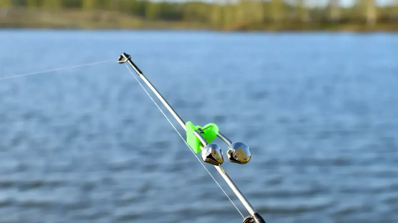 Zielony klips z dwoma dzwonkami zamontowany na wędce, na tle wody.