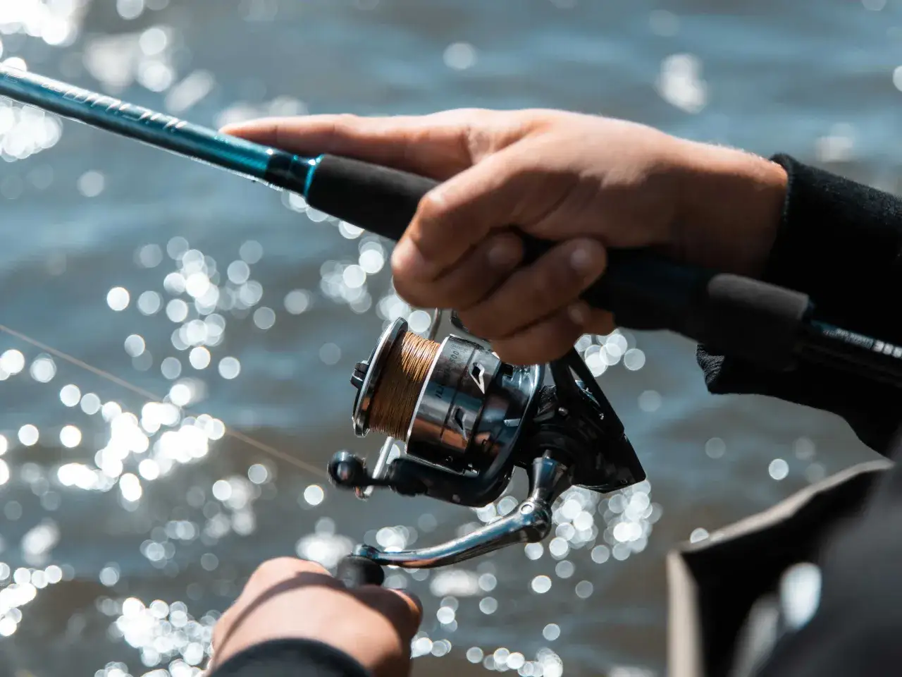 Ręce przygotowują wędkę, montując kołowrotek. Woda lśni w tle.