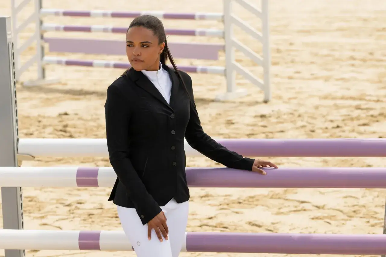 Kobieta w czarnym fraku jeździeckim i białych bryczesach stoi przy parkurze.