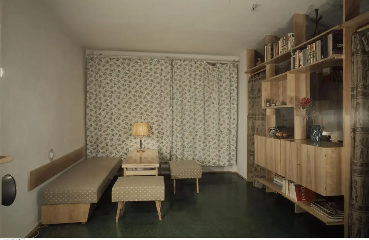 Pokój lata 80. z łóżkiem, stolikiem, pufami i regałem z książkami.