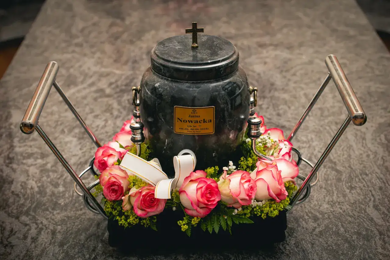 Ciemna urna z krzyżem i tabliczką, otoczona wieńcem z różowych róż i zieleni.