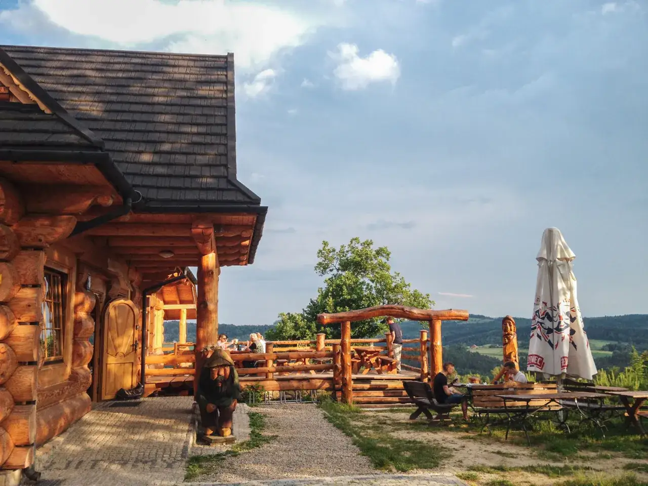 Drewniana bacówka Jamna z tarasem, gdzie goście jedzą, a obok stoi parasol. Menu bacówki Jamna kusi widokiem.