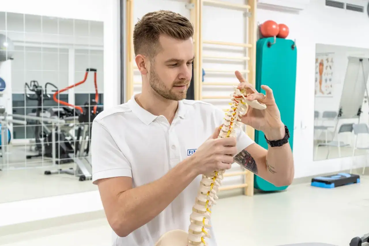 Fizjoterapeuta pokazuje model kręgosłupa, wyjaśniając, czy fizjoterapia jest trudna. W tle sprzęt rehabilitacyjny.
