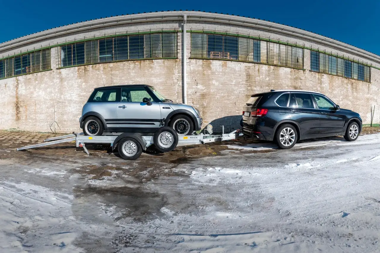 Srebrny Mini Cooper na lawecie, przygotowany do transportu. Obok stoi ciemny SUV. Mocowanie auta na lawecie jest kluczowe dla bezpiecznego przewozu.