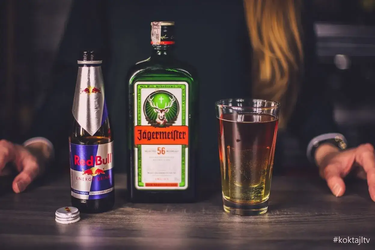 Jägermeister z czym pić? Klasycznie z Red Bullem, tworząc popularny "Jägerbomb". Na stole butelka Jägermeistera, Red Bull i szklanka z drinkiem.