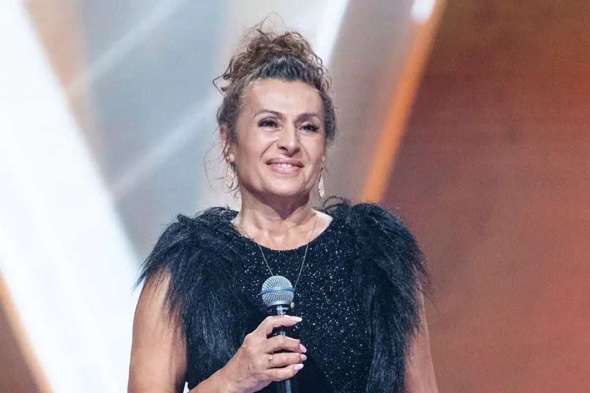 Uśmiechnięta finalistka, która mogła wygrać Voice Senior, w czarnej, błyszczącej sukni z futrzanym kołnierzem, trzyma mikrofon.