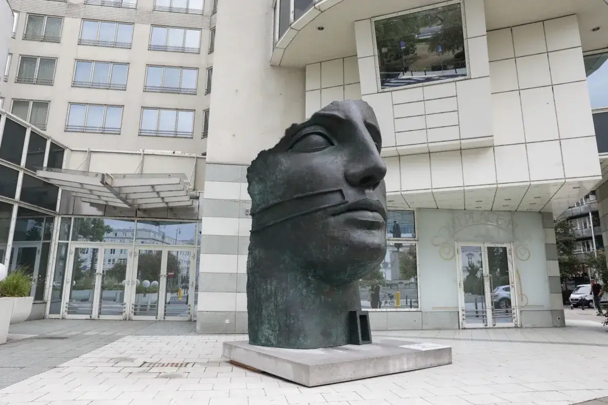 Okaleczona, ciemna rzeźba głowy z opaską na ustach, na tle nowoczesnego budynku.