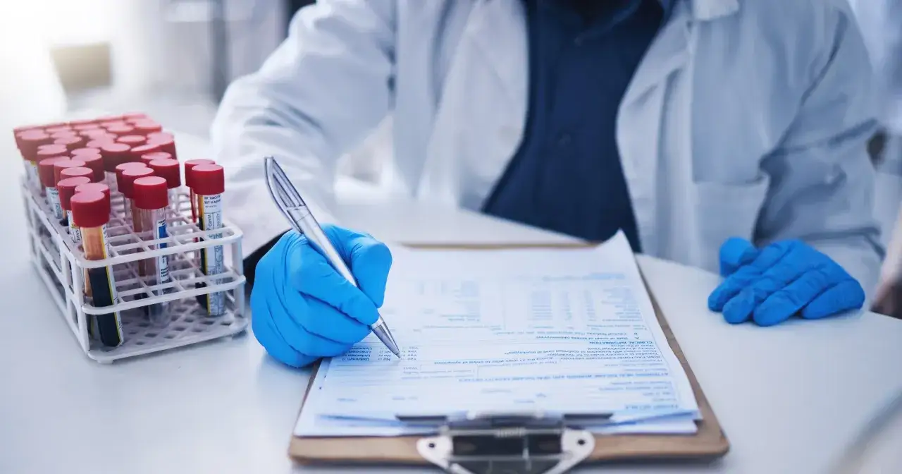 Laborant w niebieskich rękawiczkach wypełnia dokumentację medyczną obok stojaka z probówkami.