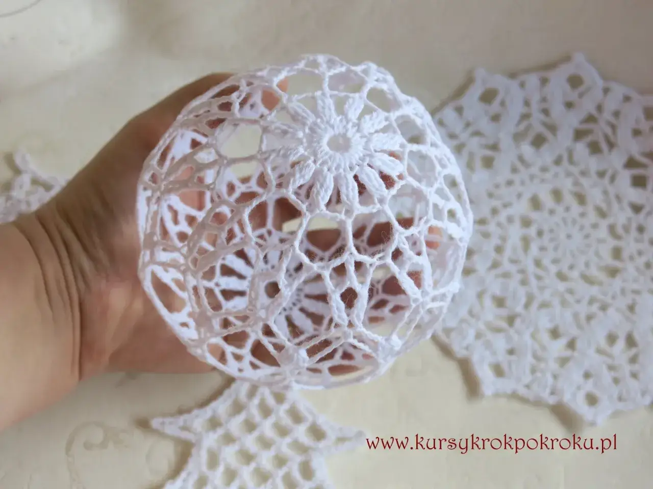 Oto jak zrobić bombkę na szydełku. Delikatna, biała, ażurowa ozdoba w dłoni, obok gotowe wzory.