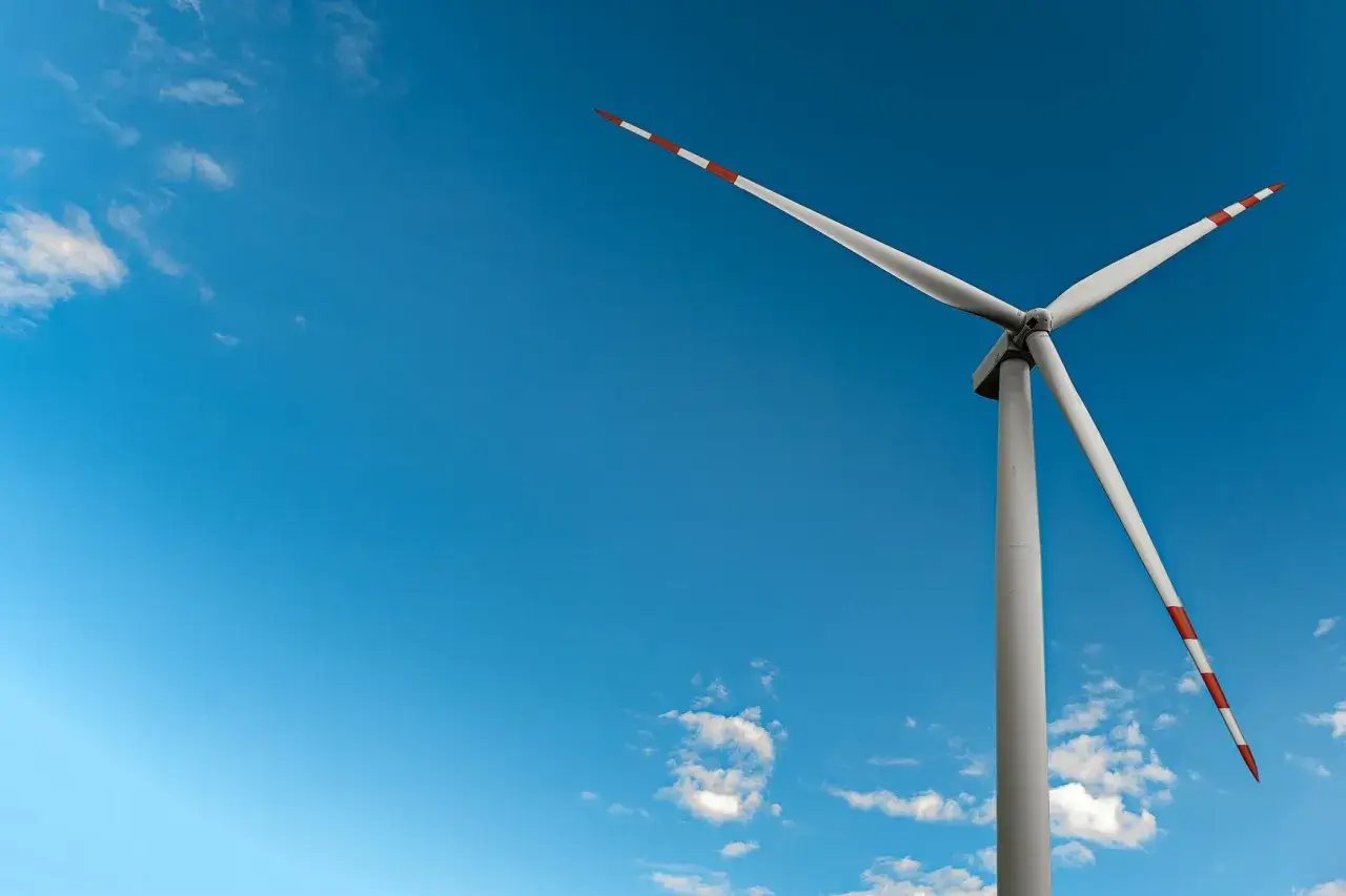 Mała elektrownia wiatrowa z białymi łopatami i czerwono-białymi końcówkami na tle błękitnego nieba z chmurami.