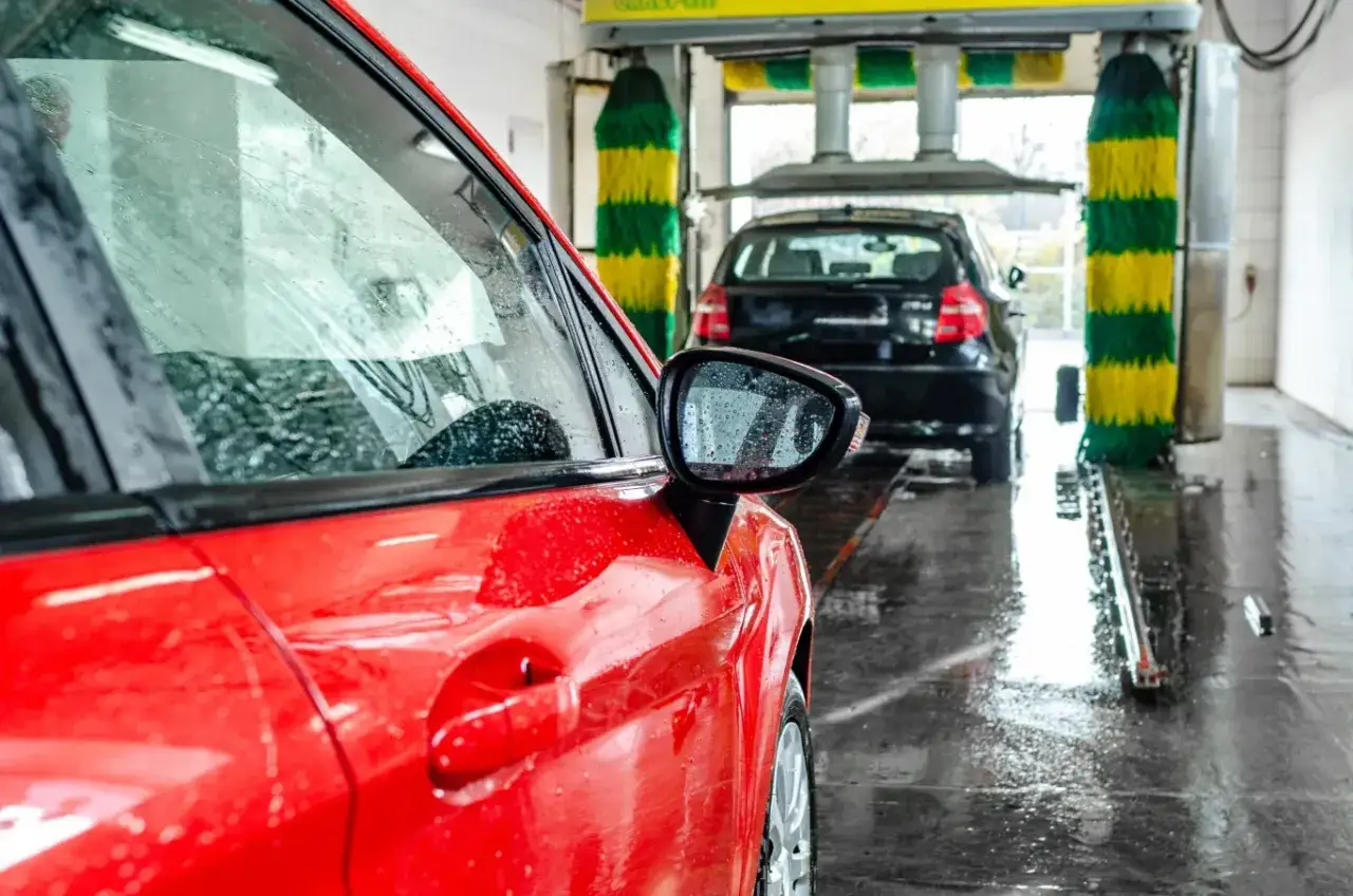 Czerwony samochód czeka na mycie w myjni automatycznej. W tle czarny pojazd i kolorowe szczotki. Wybierz najlepszy shell myjnia programy.