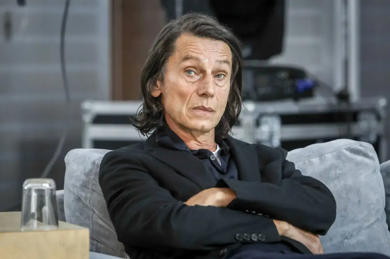 Krzysztof Warlikowski, reżyser teatralny, w czarnym garniturze, siedzi na kanapie. Jego życie prywatne budzi zainteresowanie.