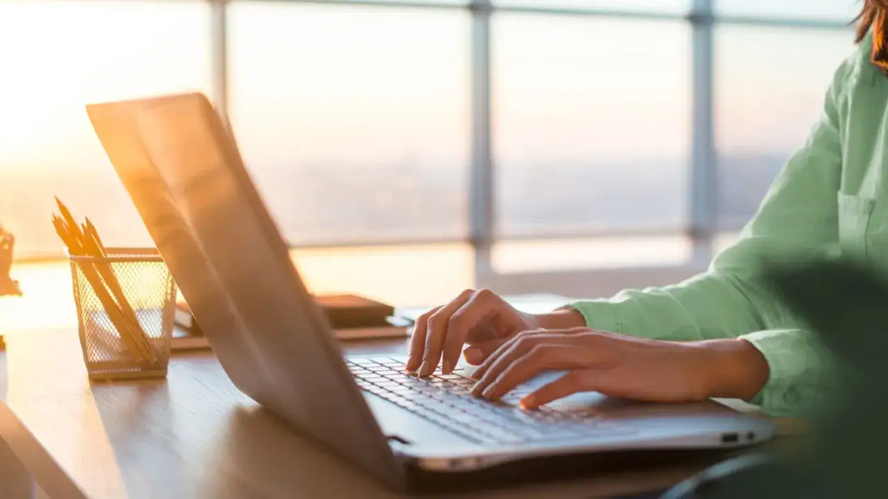 Dłonie piszące na klawiaturze laptopa w świetle zachodzącego słońca.