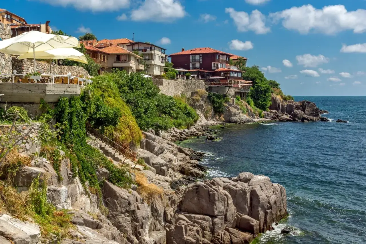 Skaliste wybrzeże z domami na klifie, restauracja z parasolami. Szukasz miejsca, gdzie nad morzem jest najtaniej?