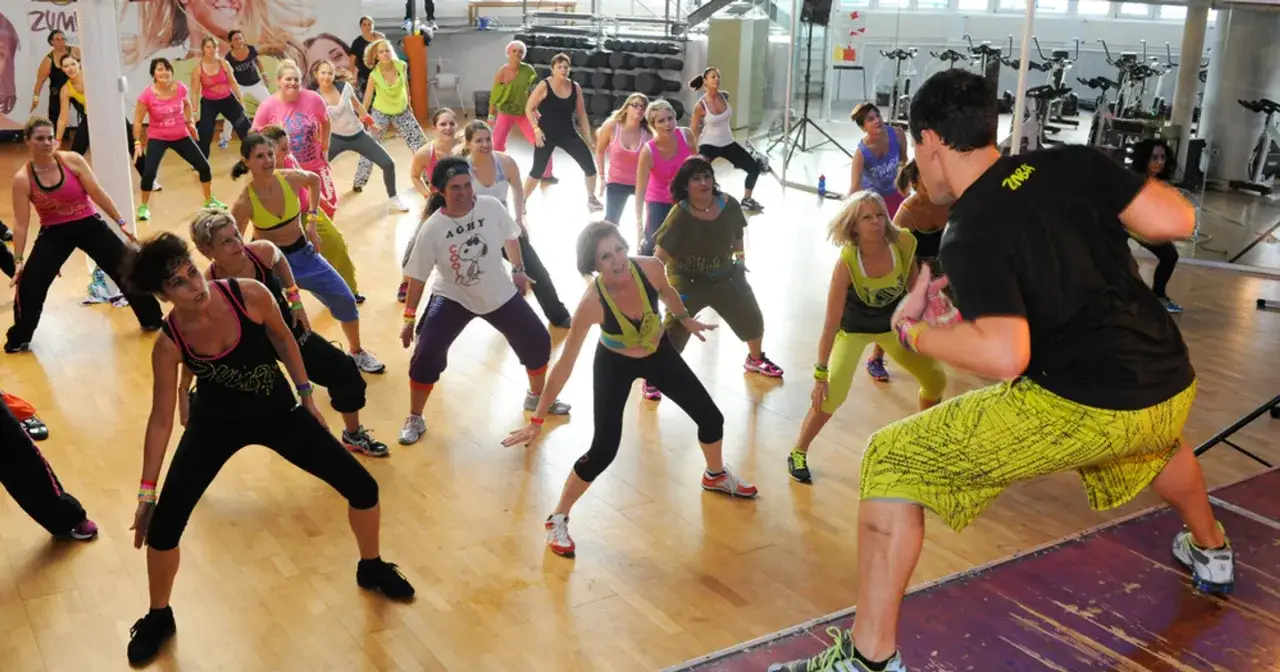 Grupa kobiet ćwiczy podstawowe kroki zumby w sali gimnastycznej, naśladując ruchy instruktora.