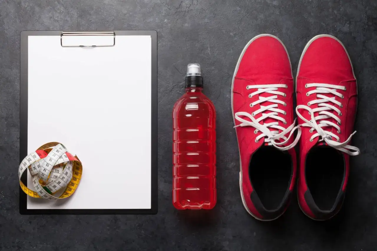 Tabela węglowodanów w produktach, miarka krawiecka, czerwony napój izotoniczny i buty sportowe.
