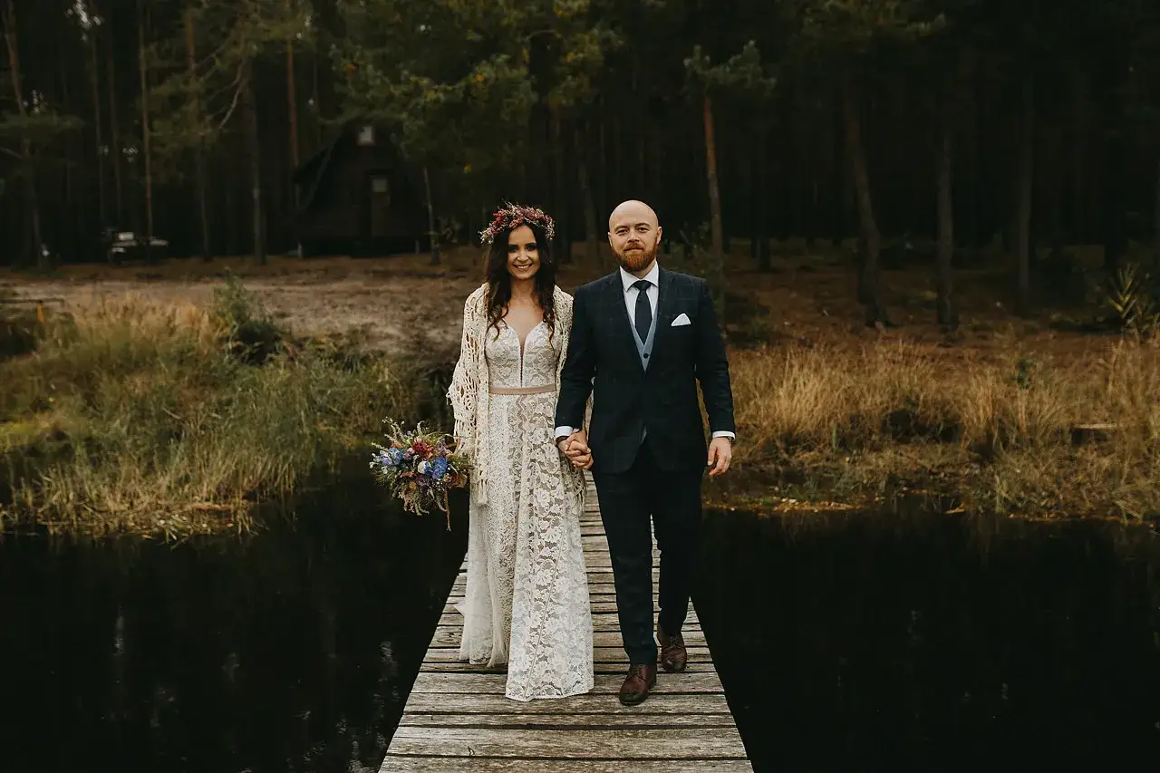 Para idzie przez las, ona w koronkowej sukni i wianku, on w garniturze. Ich suknia ślubna boho rustykalna idealnie pasuje do leśnej scenerii.
