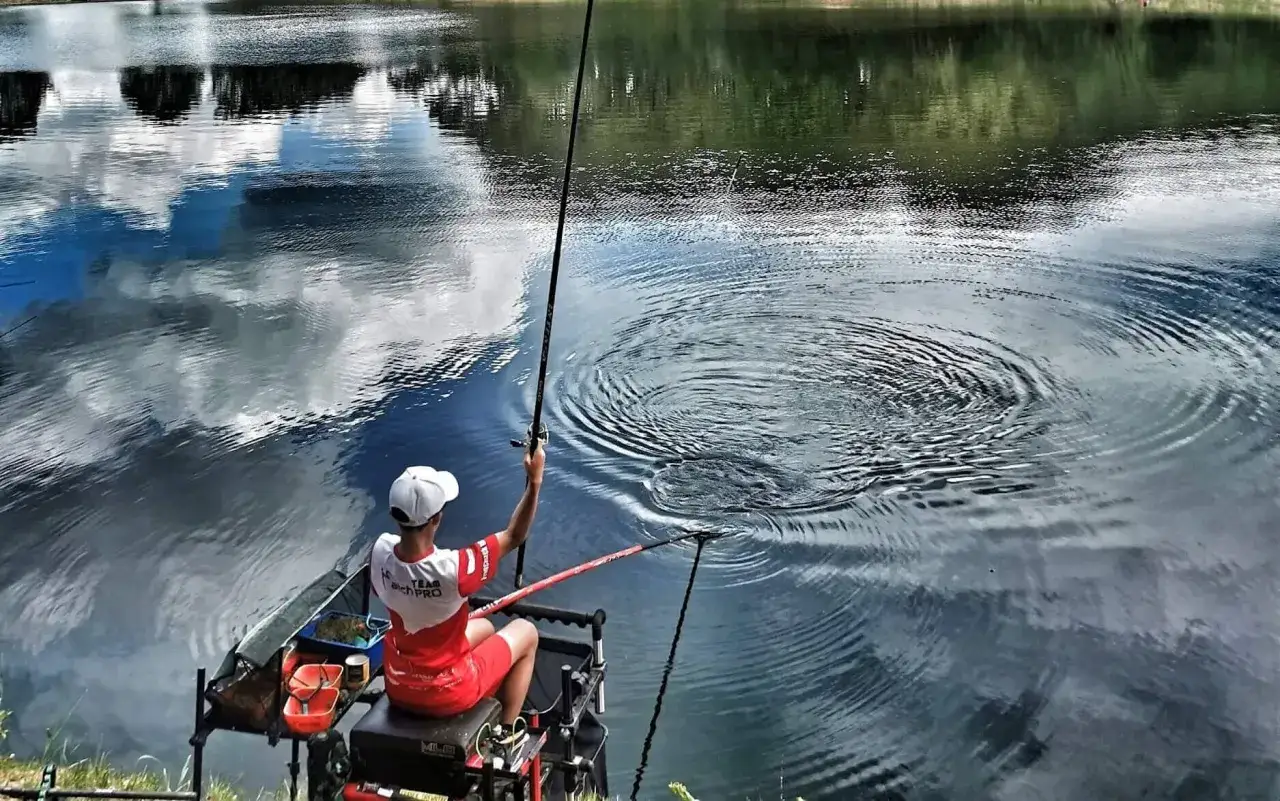 Młody wędkarz z wędką spławikową czeka na branie. Woda faluje, odbijając chmury.