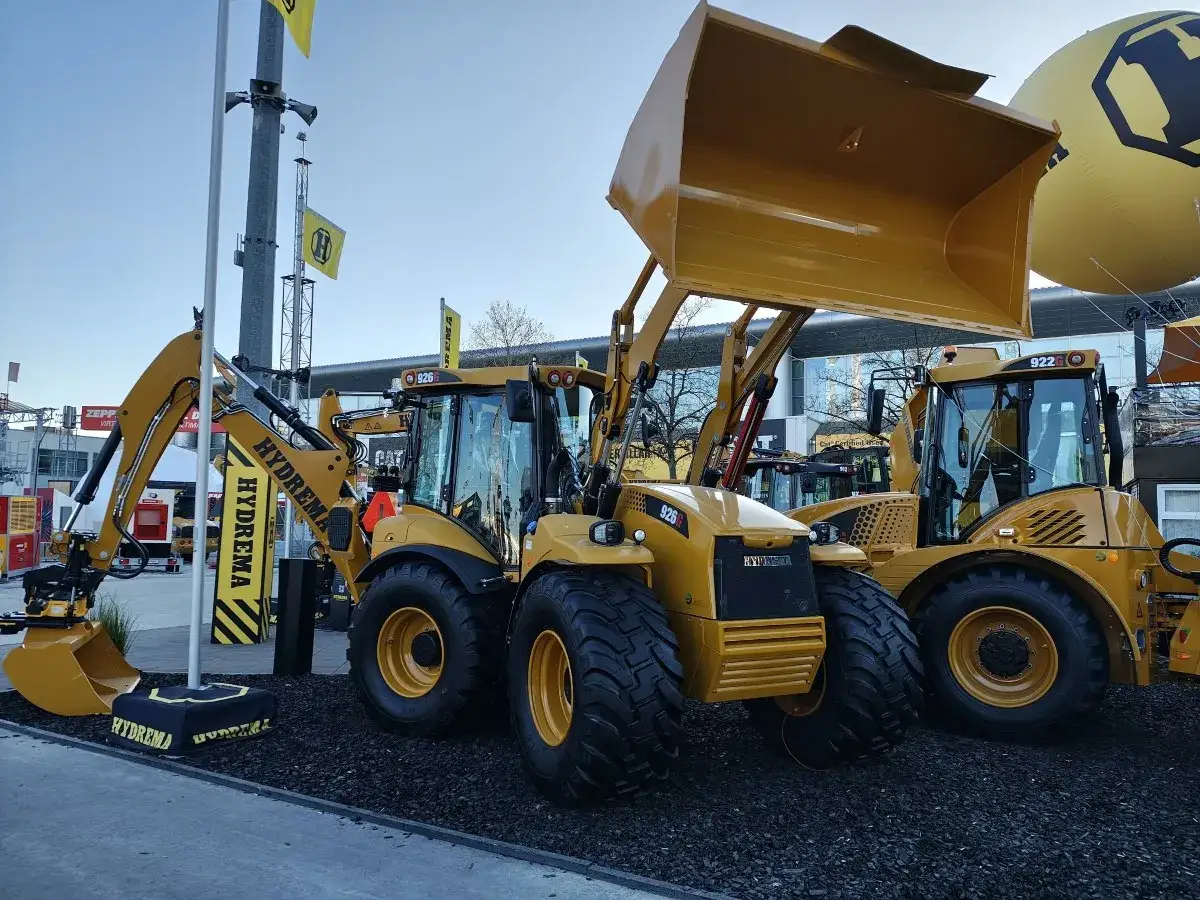 Dwie żółte koparko-ładowarki Hydrema gotowe do pracy. Jedna z nich ma podniesioną łyżkę, gotową do załadunku.