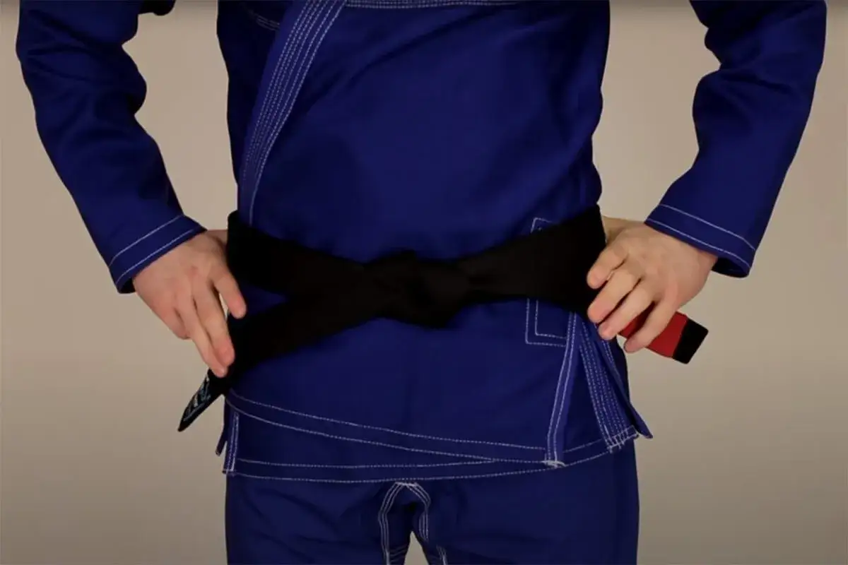 Osoba w niebieskim gi demonstruje wiązanie pasa bjj. Czarny pas jest ciasno zawiązany na wysokości talii.