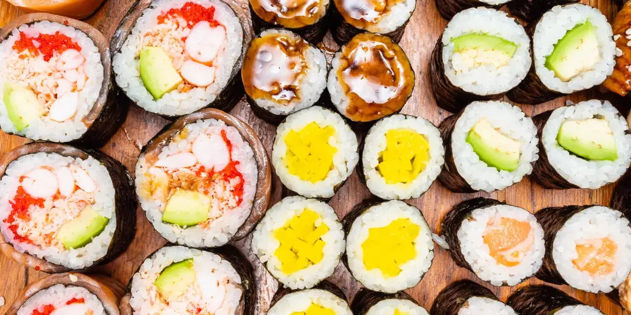 Różnorodne rolki sushi: grubsze futomaki z krewetkami i awokado, cieńsze hosomaki z łososiem, awokado, żółtą rzodkwią i polane sosem.
