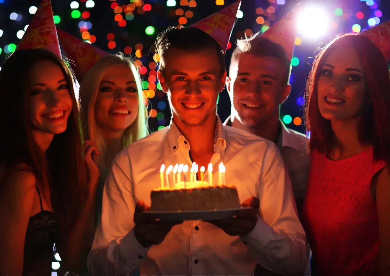 Uśmiechnięty mężczyzna z tortem i płonącymi świeczkami, otoczony przyjaciółmi na imprezie. 100 lat wszystkiego najlepszego z okazji urodzin!