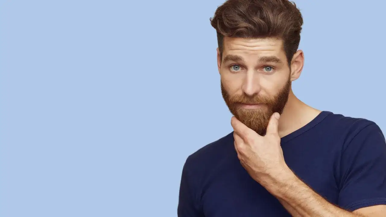 Mężczyzna z rudą brodą i niebieskimi oczami dotyka brody na jasnoniebieskim tle.