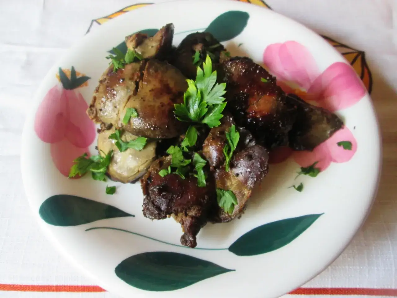 Usmażona wątróbka drobiowa, udekorowana natką pietruszki, na talerzu z kwiatowym wzorem. Idealna do przygotowania, jak oczyścić wątróbkę drobiową.