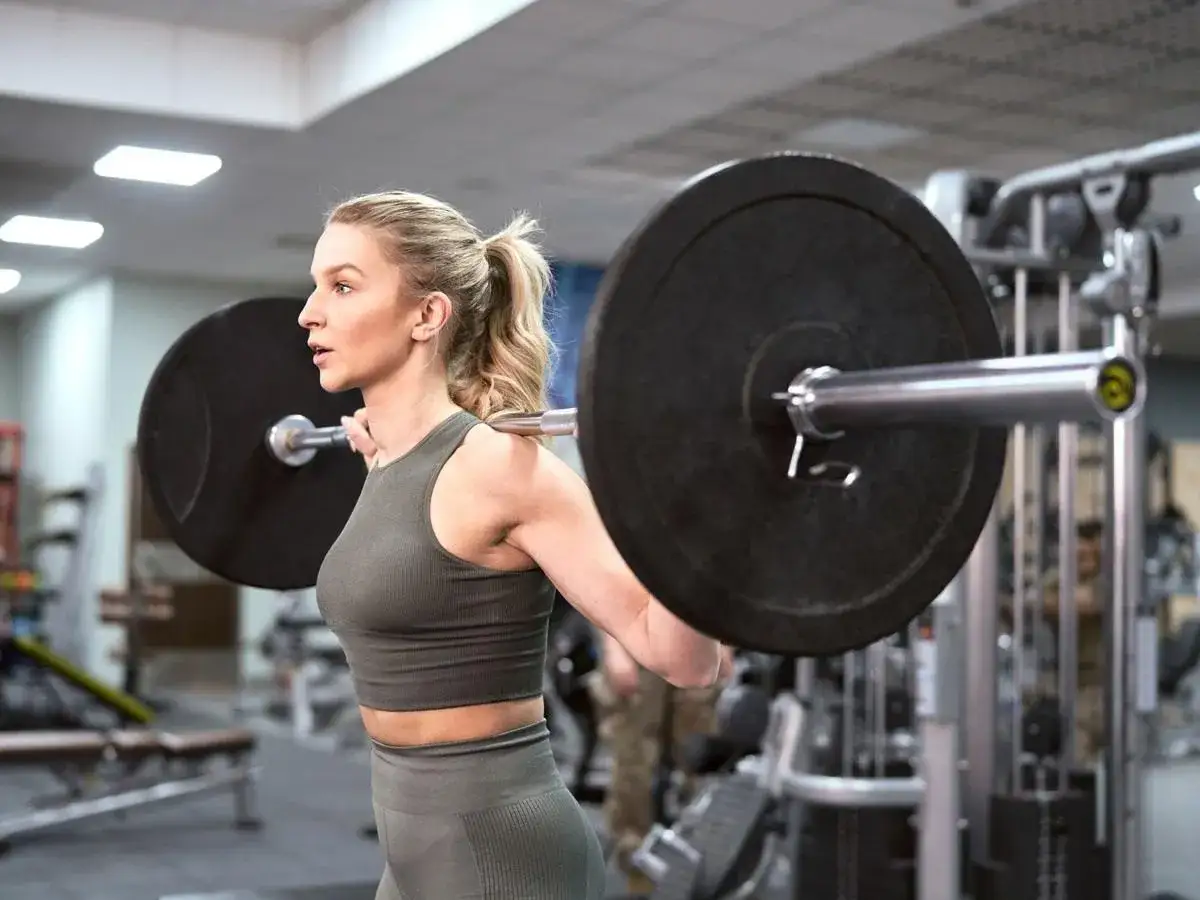 Kobieta z blond kucykiem ćwiczy z ciężarem na sztandze w siłowni.