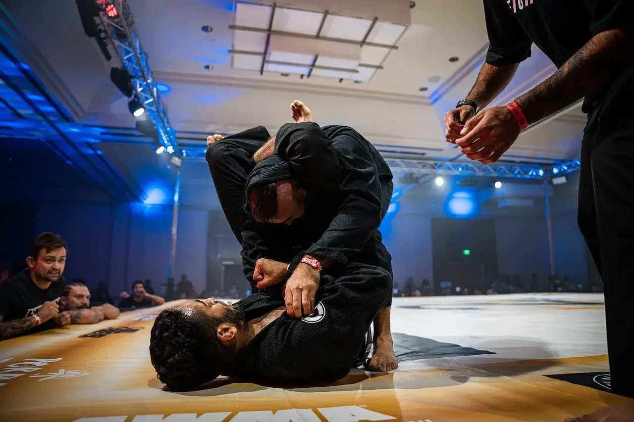 Dwóch mężczyzn w czarnych kimonach walczy w parterze. Intensywna walka jiu jitsu, z widownią w tle.