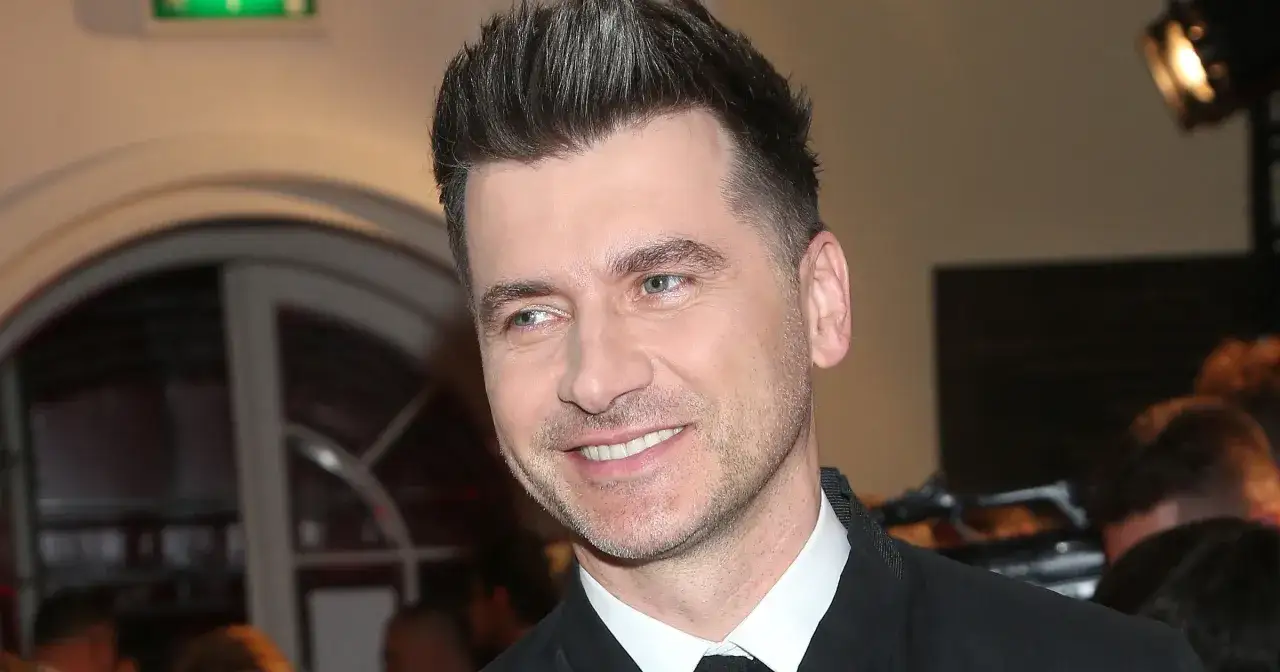 Uśmiechnięty Tomasz Kammel, jego wiek nie jest widoczny, ale jego stylizacja i fryzura sugerują, że jest w sile wieku.