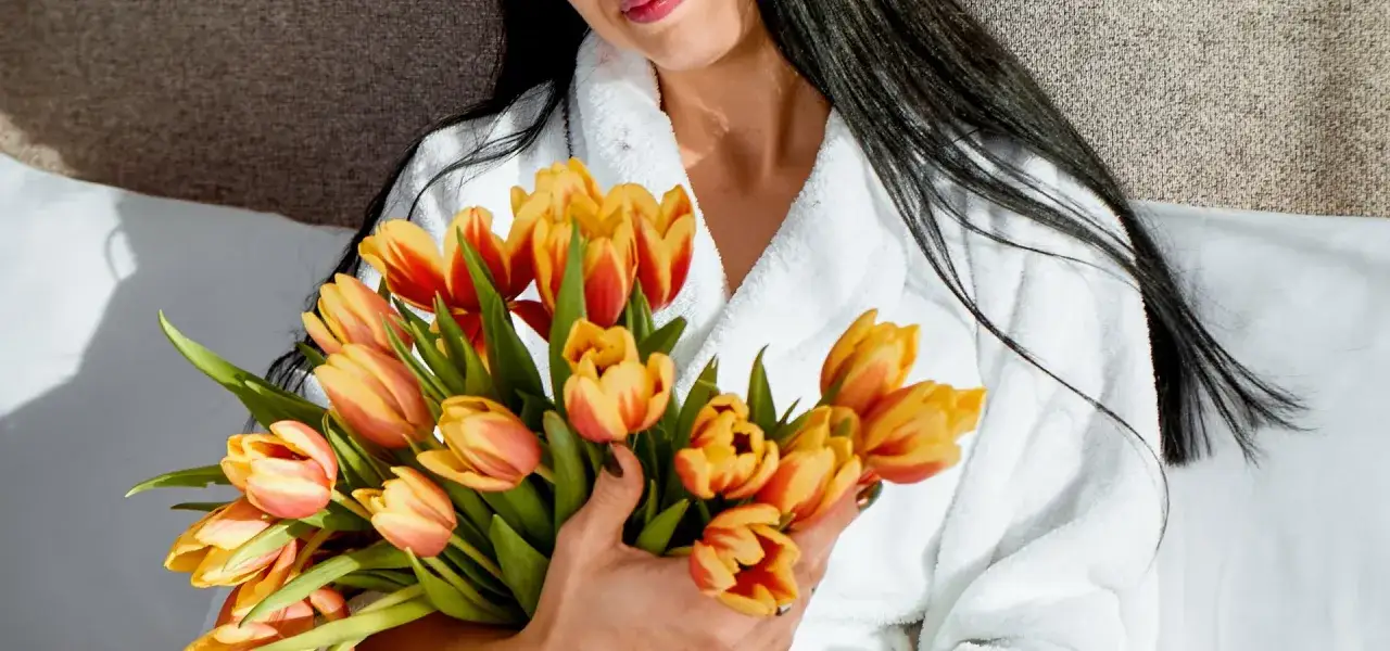 Kobieta w szlafroku trzyma bukiet żółto-czerwonych tulipanów. Idealny prezent na Dzień Kobiet, by zacząć wspaniały weekend.