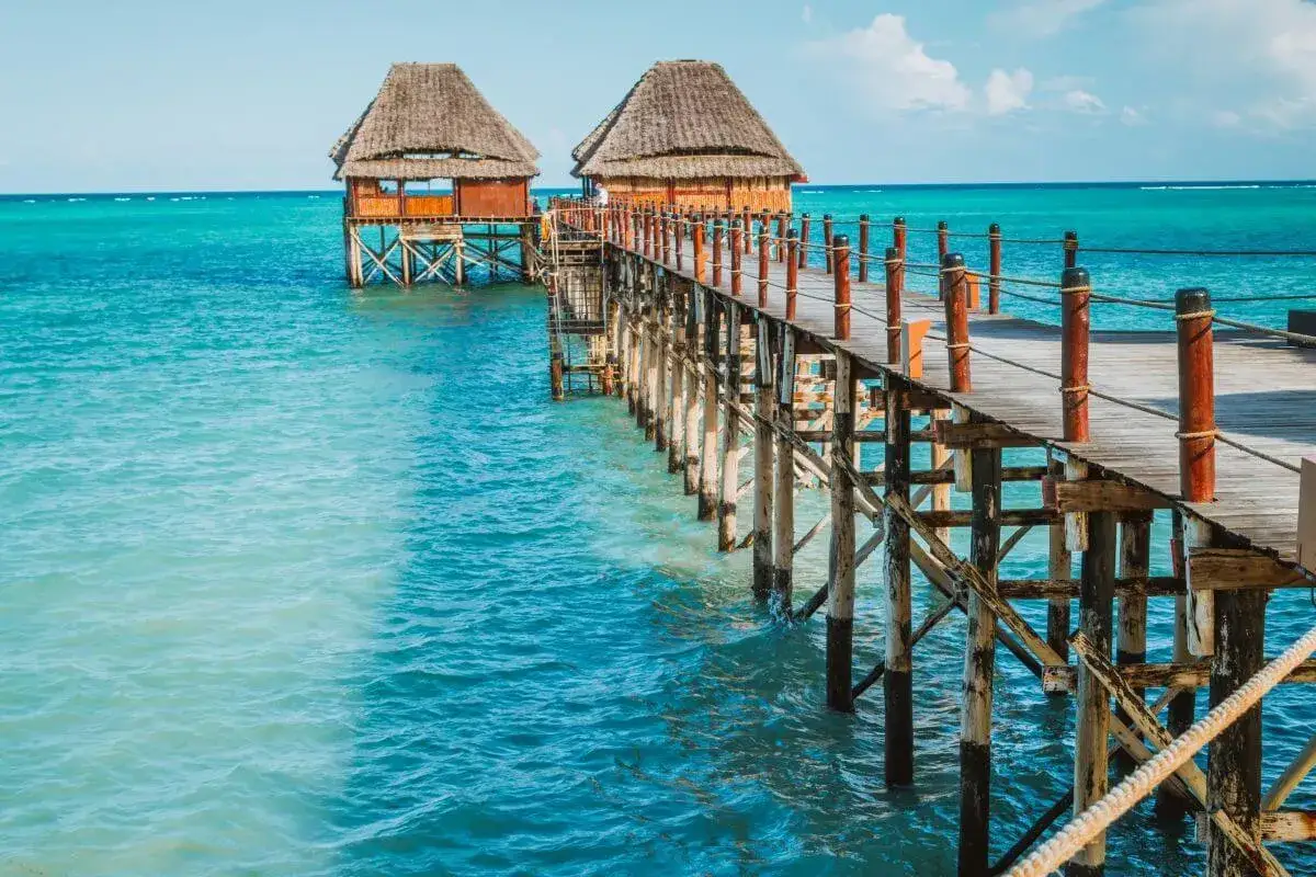Drewniany pomost prowadzi do chat na palach nad turkusową wodą. Zanzibar, pora deszczowa, ale słońce świeci.