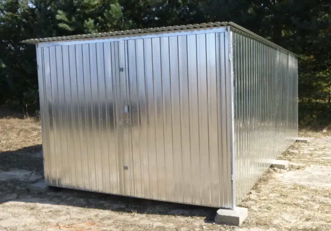 Nowy garaż blaszany z dwuskrzydłowymi drzwiami, stojący na betonowych bloczkach, na tle drzew.