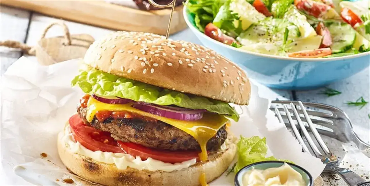 Soczysty burger z grilla z topniejącym serem, chrupiącą sałatą, pomidorem i czerwoną cebulą, podany z sałatką ziemniaczaną.
