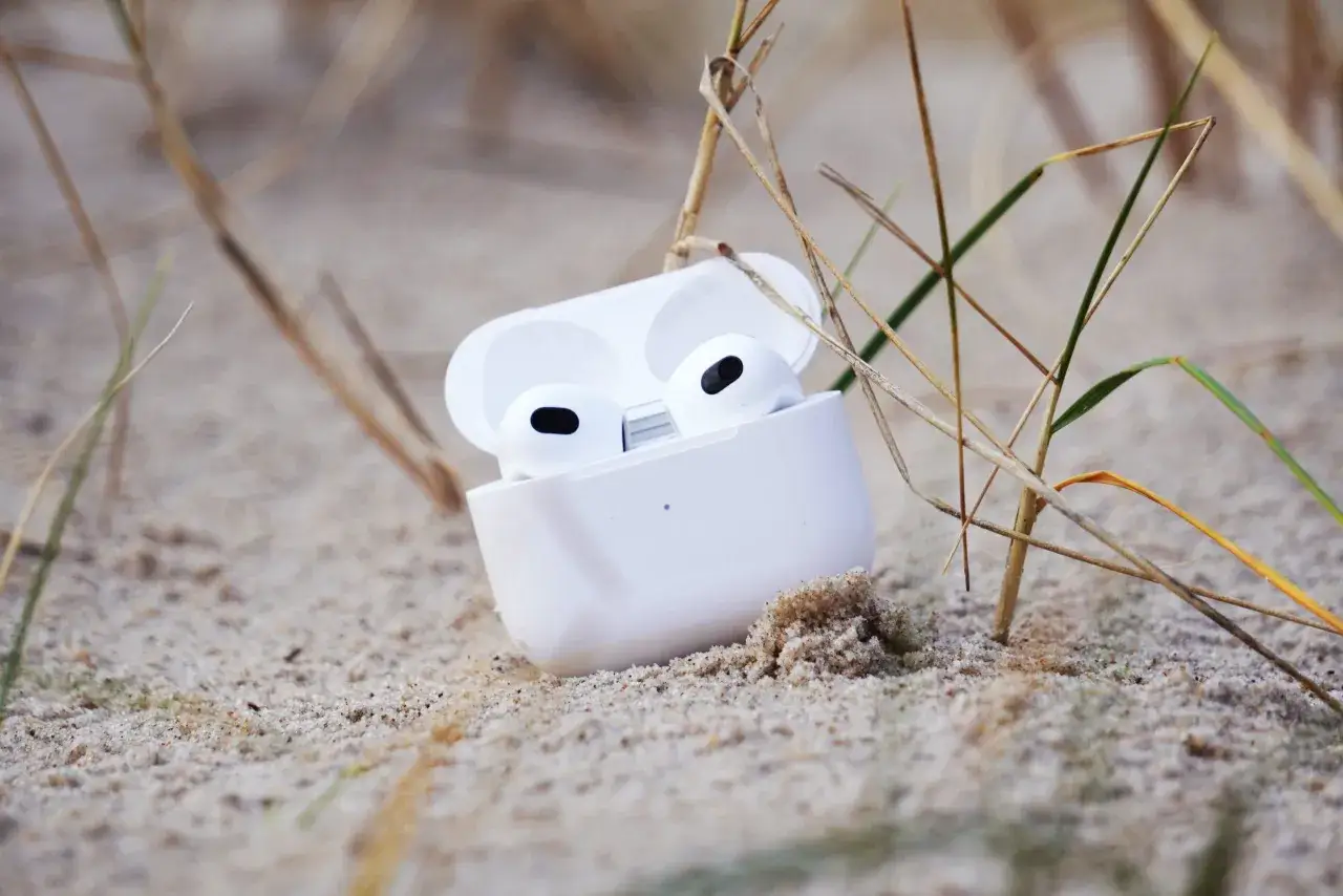 Weiße AirPods im geöffneten Ladecase liegen im Sand, umgeben von Gräsern.