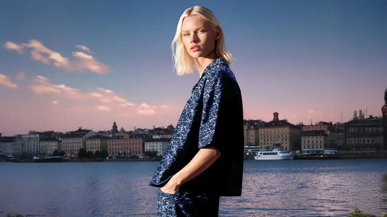 Modelka w granatowym wzorzystym ubraniu na tle miasta nad wodą o zachodzie słońca.