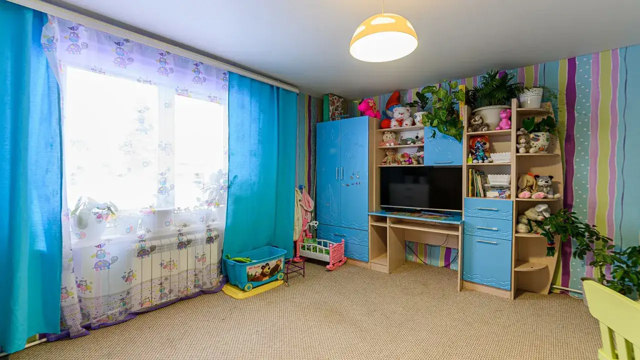 Pokój dziecięcy w stylu lat 90. z niebieskimi meblami, pluszakami i kolorowymi zasłonami.