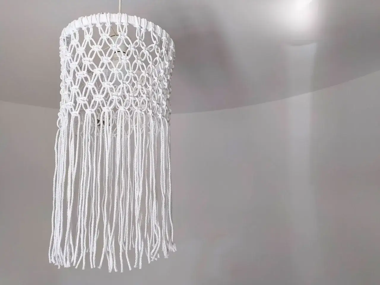 Oto jak zrobić abażur ze sznurka jutowego: ręcznie robiony, biały, makramowy klosz z frędzlami, wiszący na suficie.